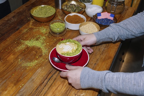 A delightful cup of specialty coffee crafted with intricate pistachio latte art on a wooden table. Surrounding ingredients include spices and seeds, showcasing artisanal coffee preparation