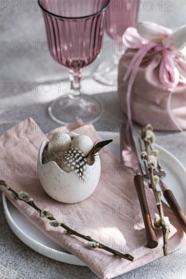 Soft pastel pink easter table setting decor. A white ceramic egg holder, adorned with subtle feather, is surrounded by pink glassware and a matching fabric pouch and pussy willow branches on a textured grey background