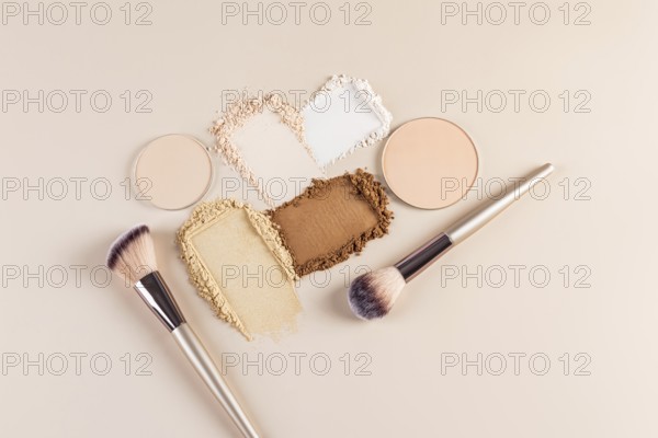 Top view of compact and loose face powders scattered artistically next to makeup brushes on a beige background. The image showcases makeup textures and tools, ideal for beauty concepts