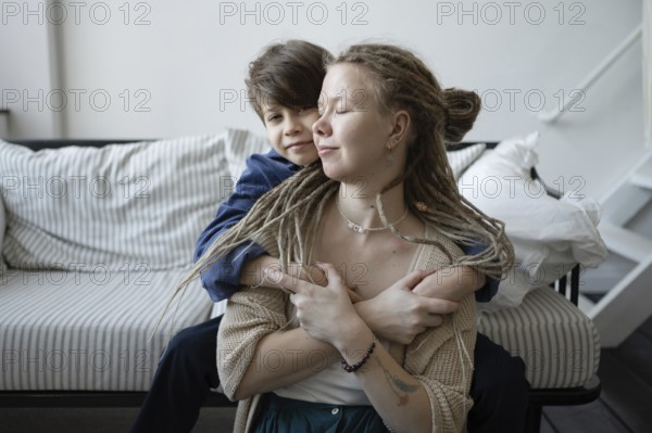 A tender moment between a mother and her son as they hug affectionately on a striped sofa The woman, with dreadlocks, closes her eyes, savoring the warmth of their connection