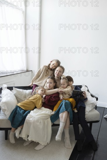 A large family sits together on a sofa in a cozy, bright living room They appear relaxed and content, surrounded by pillows, capturing a moment of togetherness