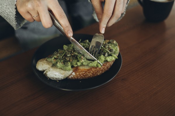 Close-up of hands using a fork and knife to slice avocado toast on a dark plate. The open-faced sandwich features fresh, creamy avocado on a slice of rustic bread