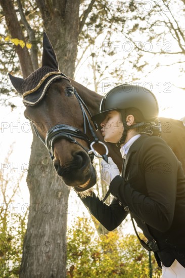 From below teenage girl, dressed in classical dressage attire, is affectionately interacting with a brown horse. She is closing her eyes and about to kiss the horse. The scene is illuminated by soft, natural light, creating a serene atmosphere