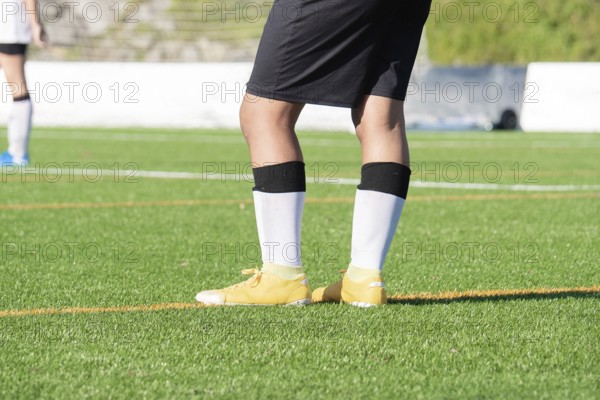A soccer player stands on a vibrant green soccer field, wearing yellow cleats, black shorts, and white socks. The background features blurred teammates, emphasizing the game's action