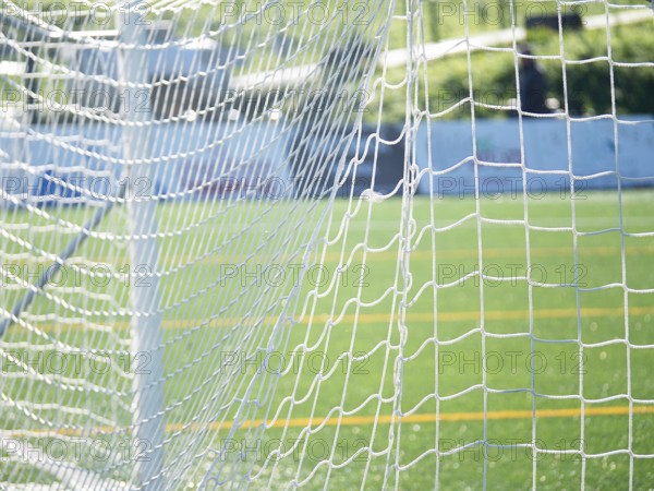 Close-up of a soccer goal net with a focus on the grid pattern under bright sunlight. Blurred green field and white lines create a vibrant, dynamic sports scene