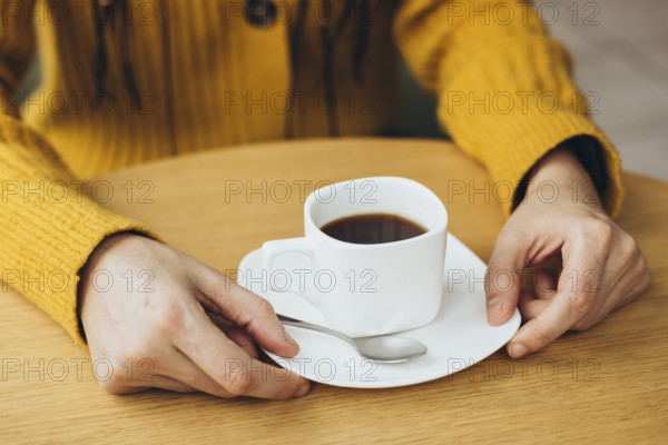 A cozy scene of a woman holding a cup of coffee on a wooden table She wears a warm yellow sweater, creating a comforting and inviting atmosphere Perfect for coffee-related themes