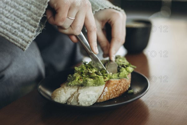 A close-up of hands slicing avocado toast topped with greens on a black plate, accompanied by a coffee cup on a wooden table. Perfect for breakfast or brunch themes