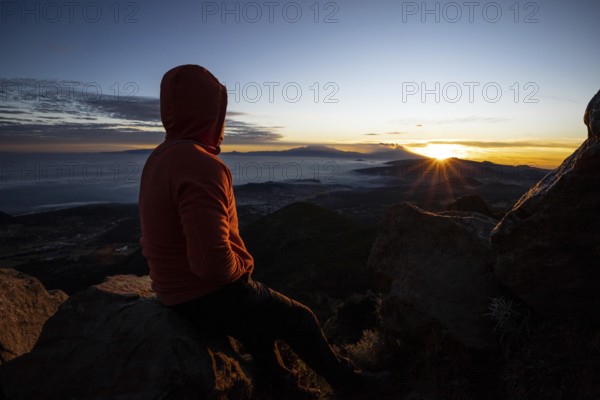Back view of an unrecognizable man sitting on a mountain peak, watching a stunning sunrise over a cloudy landscape