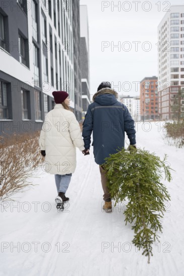 A couple walks hand in hand through a snow-covered urban area, carrying a freshly cut Christmas fir They are dressed warmly with coats and hats, capturing the holiday spirit