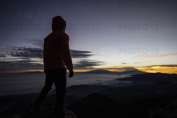 Back view of unrecognizable man standing at the summit during sunrise, overlooking distant mountains and clouds, with vibrant sky colors in the background