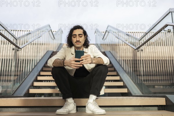 Young man sitting on outdoor stairs, casually browsing his smartphone The setting is urban and contemporary, showcasing modern lifestyle and technology engagement