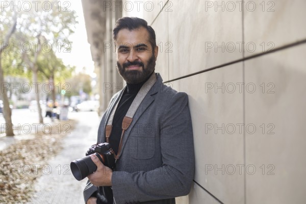 Indian photographer strolling confidently through an urban setting, showcasing a professional camera, dressed in smart winter attire, and smiling while looking at the camera