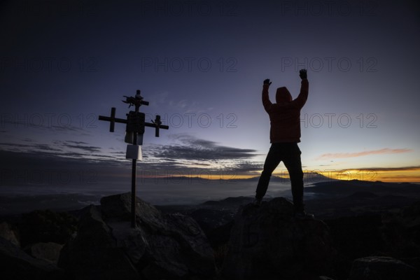 Unrecognizable man stands with arms raised near a silhouetted cross at sunrise, atop a rugged mountain peak, highlighting a moment of triumph and peace