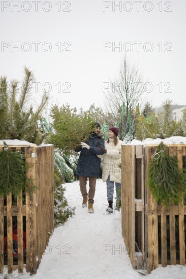 A couple strolls through a snowy Christmas fir farm, happily selecting their perfect tree Surrounded by pine trees and snow, they embrace the festive spirit
