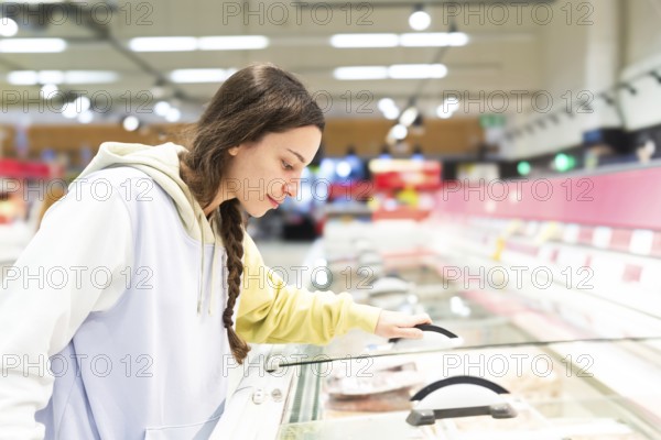 Side view of a woman is observed closely examining the products in a supermarket freezer section, apparently deciding what to buy