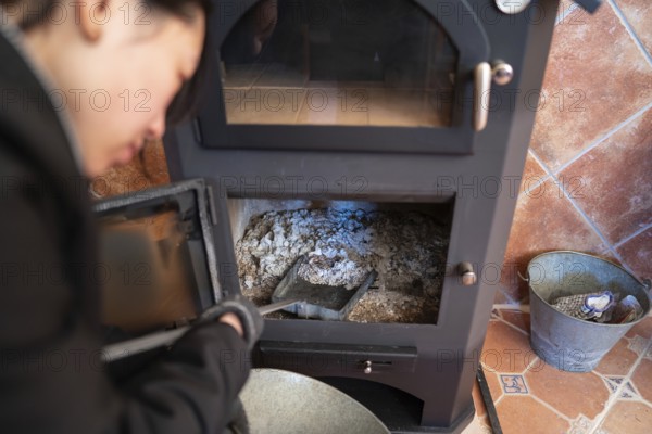 Cropped blurred Asian woman, looking down, is cleaning the ashes from a wood burning stove, holding a shovel, inside a cozy room with tiled flooring