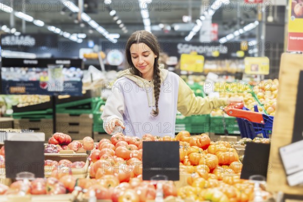 Woman in a casual hoodie shopping for fresh tomatoes in a vibrant supermarket, carefully choosing the best ones for her meal preparation
