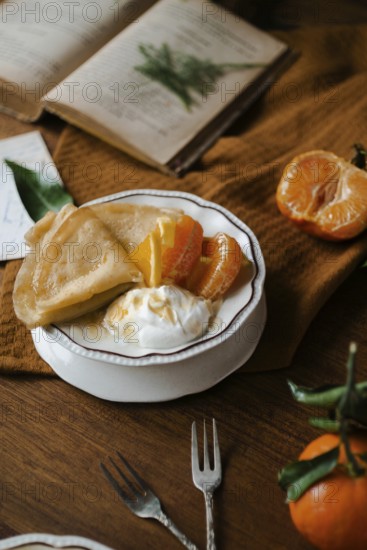 Delicious crepes filled with citrus fruits and yogurt, served on a vintage dish beside a rustic book and some fresh mandarins on a wooden table