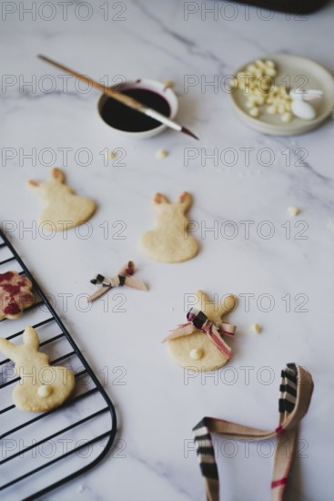 Top view of freshly baked Easter bunny cookies decorated with hibiscus tea paint, displayed on a cooling rack beside a bowl of white icing and a paintbrush dipped in hibiscus tea on a marble surface