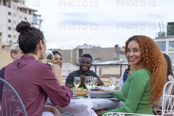 A group of friends enjoying a rooftop party, sharing drinks, food, and laughter under a clear sky They are seated around a table, engaging in conversation and celebration