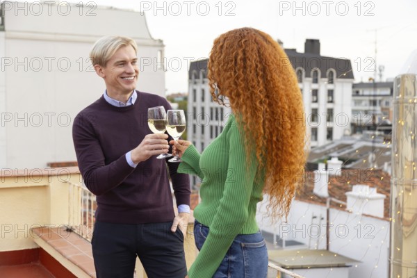 Two friends share a cheerful moment, toasting with glasses of white wine on a rooftop The urban backdrop, warm smiles, and casual attire create a lively and relaxed atmosphere