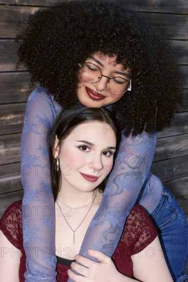 Two multiethnic Gen Z women sharing a close embrace, displaying friendship and joy. The Hispanic woman with curly hair and glasses is hugging her caucasian friend from behind, both smiling gently towards the camera