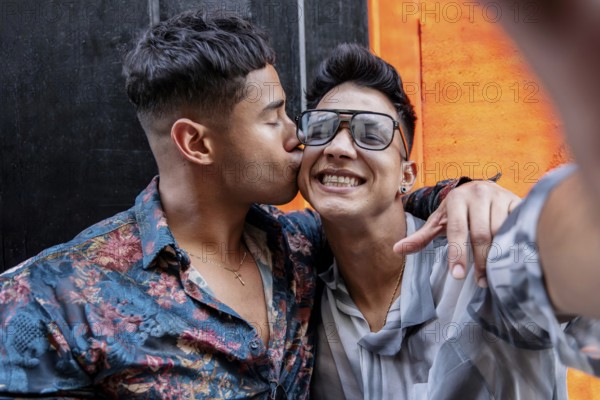 Latin gay couple taking selfie while embracing warmly. One partner kisses the other's cheek, while both are smiling under a bright orange background