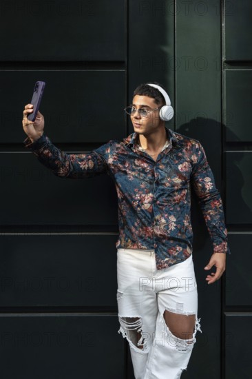Stylish man wearing floral shirt and headphones takes a selfie. He wears sunglasses and ripped jeans, standing confidently against a dark background