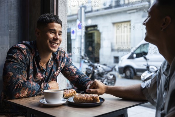 Two latin gay men hold hands, smiling and chatting over coffee and pastries in a warm, urban cafe setting, creating a scene of love and connection