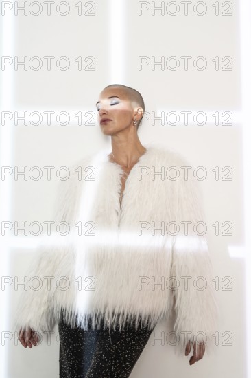 A middle-aged woman with a shaved head poses confidently, wearing a fashionable white faux fur coat and black sequined pants against a minimalist backdrop