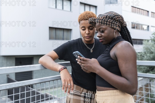 An LGBT multiethnic couple shares a tender moment outdoors, both focused on a smartphone, with one leaning on a railing and the other holding the phone. Modern urban background
