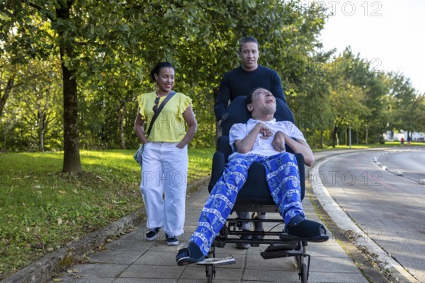 Joyful Spanish Cameroonian siblings enjoy a walk. One sibling, who has cerebral palsy, is in a wheelchair. They share smiles under the green trees on a sunny day