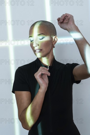 A middle-aged woman with a shaved head poses gracefully under artistic light patterns, radiating confidence and elegance in a minimalist, modern setting