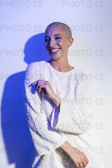 Middle-aged woman with a shaved head smiling confidently in a textured white outfit. Blue lighting casts dramatic shadows, enhancing her graceful pose