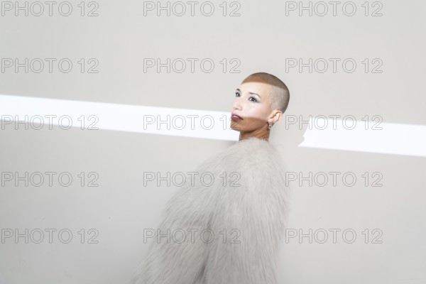 A middle-aged woman with a shaved head poses confidently, wearing a stylish furry coat against a minimalist background illuminated by a bright light strip
