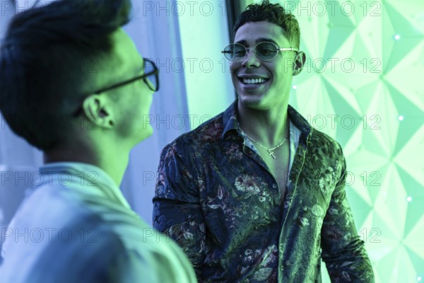 Two latin gay men smiling warmly at each other in an illuminated urban setting. Vibrant green tones create a lively atmosphere, highlighting their joyful connection and stylish attire