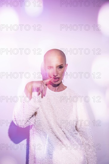 A middle-aged woman with a shaved head poses confidently, bathed in soft, bright light with lens flares creating a dreamy effect. She wears a textured white sweater