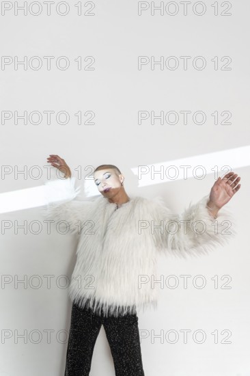 A middle-aged woman with a shaved head poses confidently in a luxurious white fur coat. The minimalist background enhances her elegant and bold fashion statement
