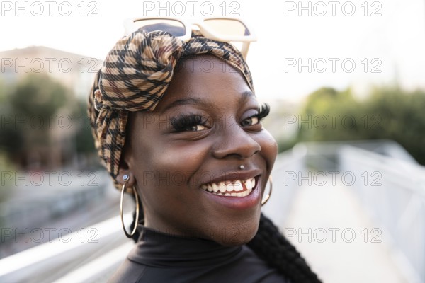 Vibrant portrait of a young black woman with a patterned headscarf and sunglasses, smiling brightly outdoors. She wears hoop earrings and exudes confidence