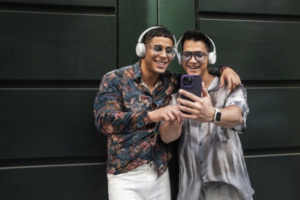 Two happy gay men wearing headphones, embracing and smiling while looking at a phone together. Their joyful expressions reflect a moment of connection and happiness