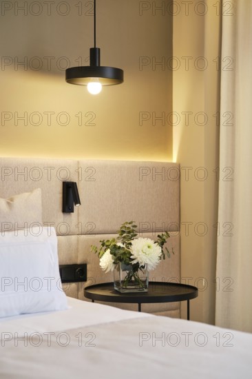 A cozy hotel room corner featuring a modern pendant lamp, cushioned headboard, and a side table with a glass vase of white flowers, exuding comfort and elegance