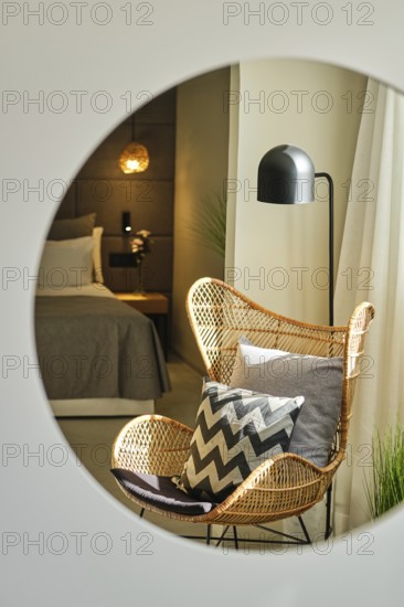 The elegant hotel room features a wicker chair with patterned cushions, a contemporary lamp and mood lighting, creating a warm and inviting atmosphere for relaxation, viewed through a mirror
