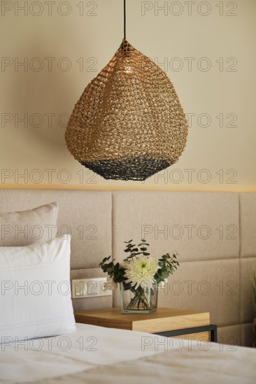 A cozy hotel room features a woven pendant lamp hanging above a wooden nightstand with a vase of white flowers. Soft lighting and neutral tones create a relaxing atmosphere
