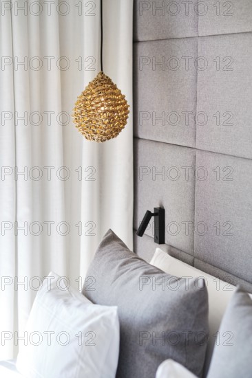 A cozy hotel room with a woven pendant lamp, plush gray pillows, and a textured gray upholstered headboard. White curtains add a touch of elegance and softness