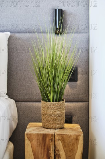 A modern hotel room detail featuring a wooden bedside table with a woven plant pot. The gray upholstered headboard adds a touch of elegance and comfort