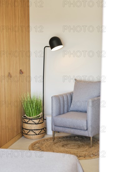 A cozy hotel room corner featuring a gray armchair, woven basket with green plant, and a sleek floor lamp. Modern wooden paneling adds warmth and style