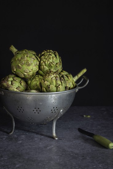 A collection of fresh artichokes sits in a rustic metal colander on a dark counter, accompanied by a small knife. The scene highlights texture and contrast