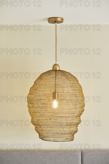 A stylish woven pendant light hangs from a hotel room ceiling, casting a warm glow. The beige color complements the room's serene and cozy atmosphere