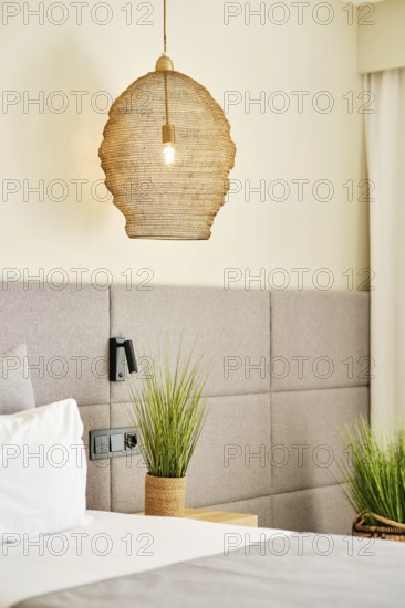 A cozy hotel room featuring a woven pendant lamp, soft gray headboard, and lush green plants. The neutral palette and natural textures create a calming and modern atmosphere