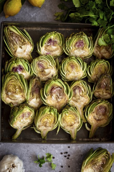 Top view of fresh artichoke halves on a baking tray, surrounded by lemons, herbs, and garlic, creating a vibrant culinary display ready for cooking